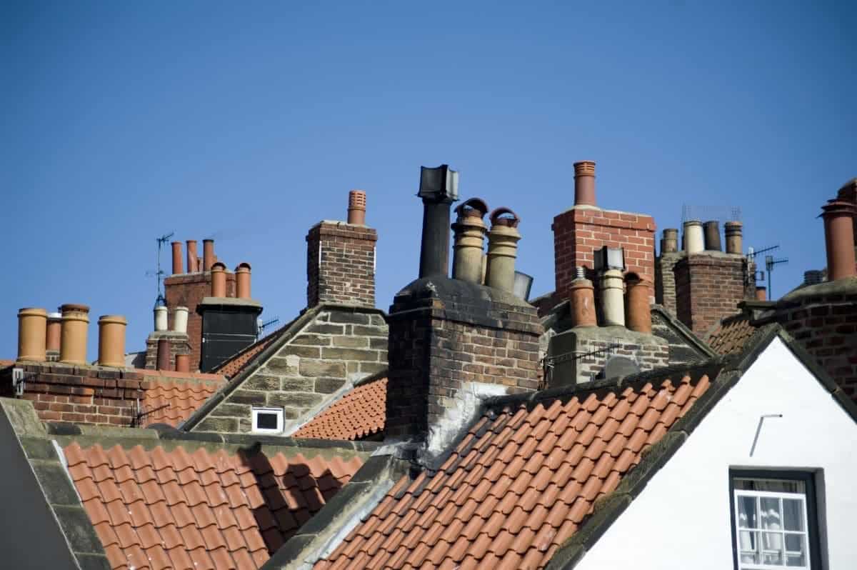 Rooftops with multiple brick chimneys and red clay tiles are set against a clear blue sky. Some roofs have white trim, and the chimneys vary in size and shape, showcasing the need for expert Chimney Repair Providence County, RI.