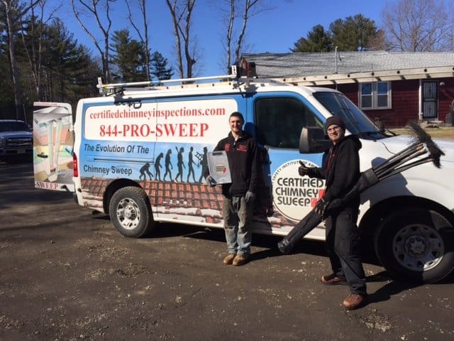 Two chimney sweeps stand next to a white "Certified Chimney Sweep" van, holding chimney cleaning tools, on a sunny day in front of a house and trees—your trusted experts for Chimney Cleaning Providence County, RI.