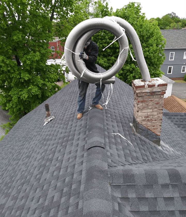 A person stands on a gray shingled rooftop in RI, holding a large coiled flexible duct near a brick chimney, preparing for what appears to be chimney or roof work. Lush green trees and neighboring houses are visible in the background.