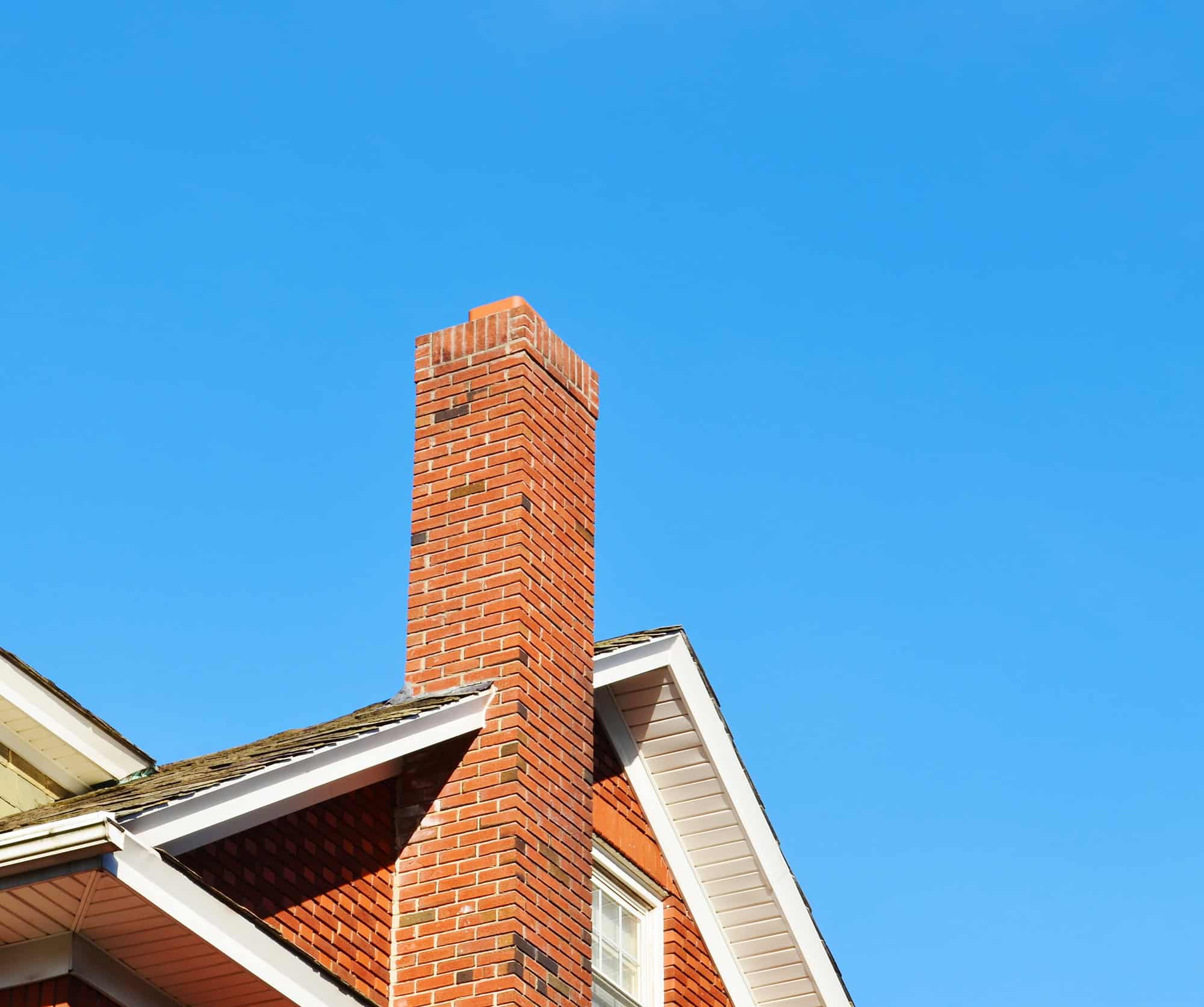 A tall red brick chimney extends above the roof of a house with white trim, set against a clear blue sky—an ideal example of homes in RI that may benefit from expert Chimney Repair Providence County services.