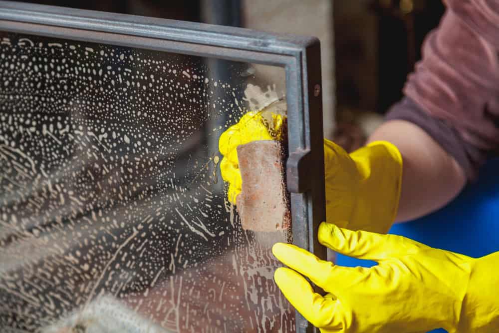 A person wearing yellow rubber gloves is cleaning a glass oven door with a sponge. The glass is covered in soapy bubbles as they scrub, similar to the care taken during a Chimney Sweep Providence County service in RI.