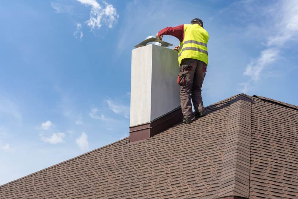 A worker in a yellow safety vest stands on a sloped roof in Providence County, RI, installing or inspecting a white chimney under a blue sky with some clouds.