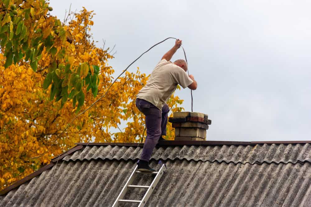 A person stands on a ladder leaning on a rooftop, cleaning or inspecting a brick chimney with a long tool—possibly as part of Chimney Repair Providence County, RI. Yellow autumn leaves appear in the background under a cloudy sky.