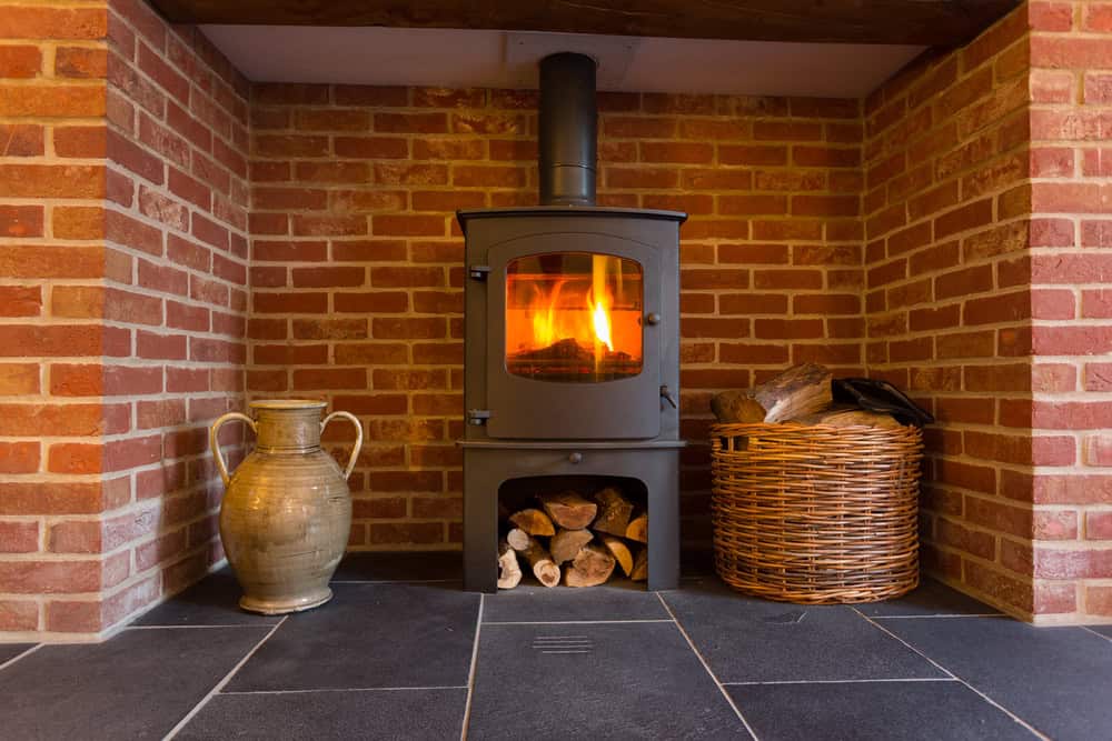 A black wood-burning stove with a fire inside sits against a red brick wall, flanked by a ceramic jug and a wicker basket of firewood on the dark tiled floor—perfect for cozy RI nights or after Chimney Cleaning Providence County services.