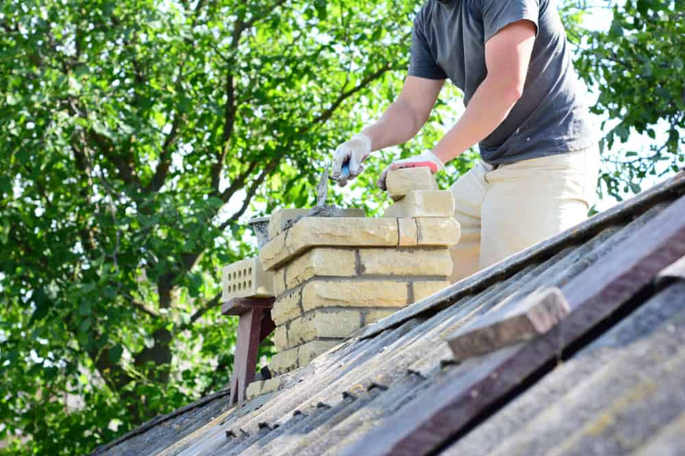 A person wearing gloves repairs a brick chimney on a sloped roof in RI, surrounded by green leafy trees in the background.