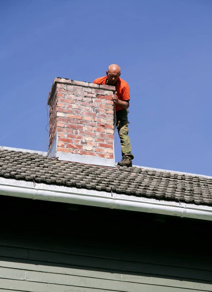 A man in an orange shirt stands on a tiled rooftop, inspecting a red brick chimney under a clear blue sky—an example of expert Chimney Repair Providence County, RI.