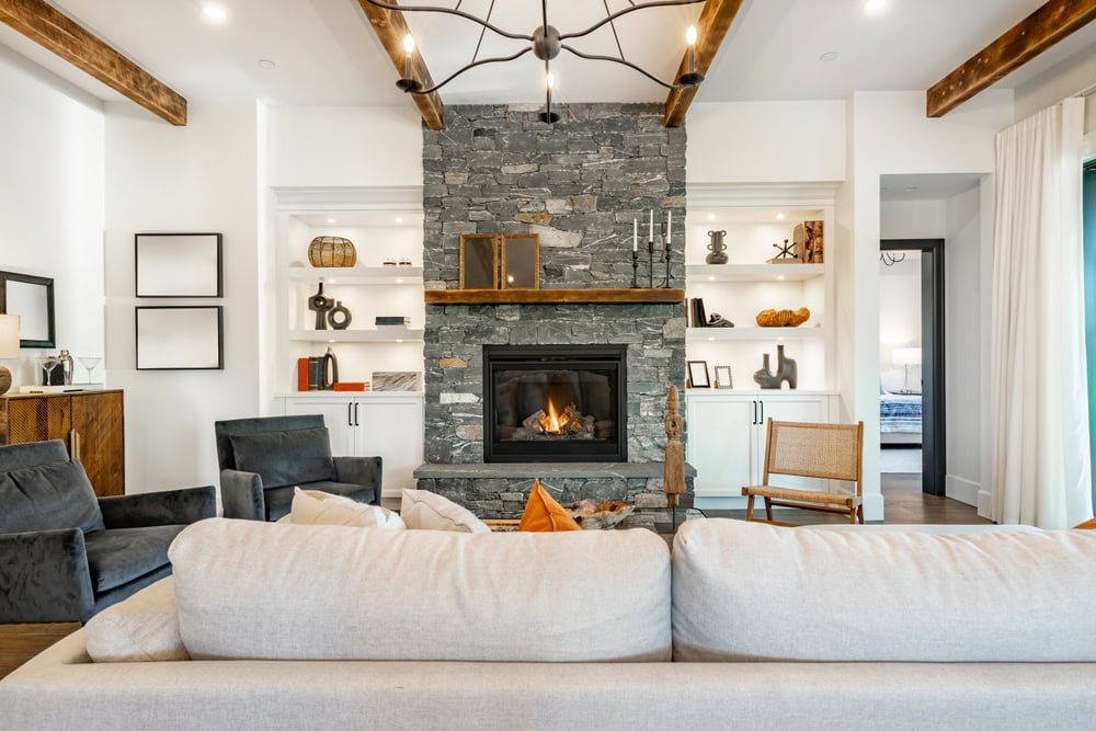 Modern living room with exposed wood beams, a stone fireplace maintained by Chimney Sweep Providence County, cozy seating, built-in shelves displaying decor, and a large window with white curtains allowing natural light into the space.