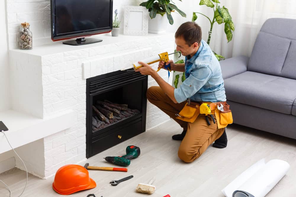 A person wearing a tool belt kneels in front of a modern white fireplace in RI, applying sealant with a caulking gun. Tools, a hard hat, and a tape measure are scattered on the light-colored floor, ready for Chimney Cleaning Providence County, RI.