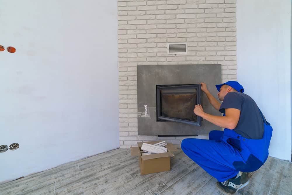 A person in blue overalls and a cap is installing or inspecting a fireplace door in a modern room with white brick walls and gray flooring—perfect for anyone seeking Chimney Sweep Providence County, RI services. A box of tools sits nearby.