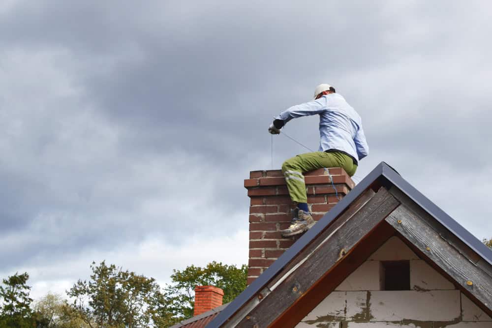 A person wearing a cap and gloves is performing Chimney Cleaning Providence County on a brick chimney atop a rooftop under a cloudy sky, with trees visible in the background.