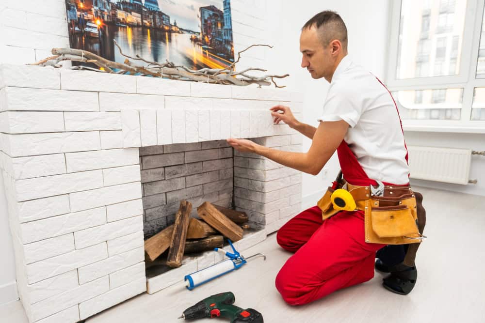 A man in red overalls and a tool belt installs white decorative panels on a fireplace. Tools, wood logs, and a painting are nearby in a bright, modern room with large windows—ideal for Chimney Repair Providence County, RI.
