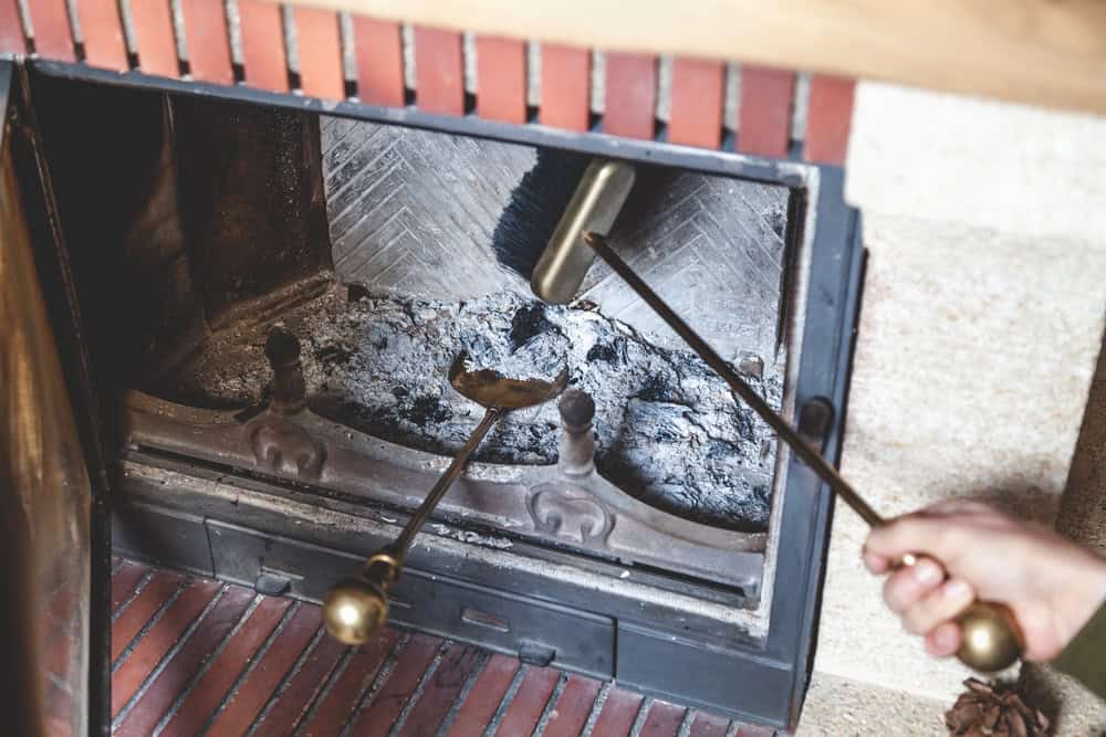 A person uses fireplace tools to clean ashes from a brick fireplace in RI. One hand holds a brush sweeping inside, and the other holds a poker. The fireplace contains metal andiron supports and leftover ashes.