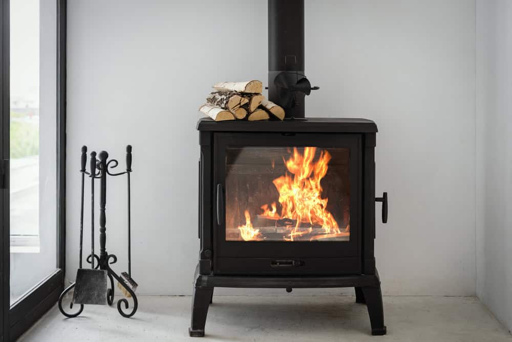 A black wood-burning stove with a fire inside sits in a corner next to fireplace tools and a stack of cut firewood on top. The room has white walls and a glass door nearby.