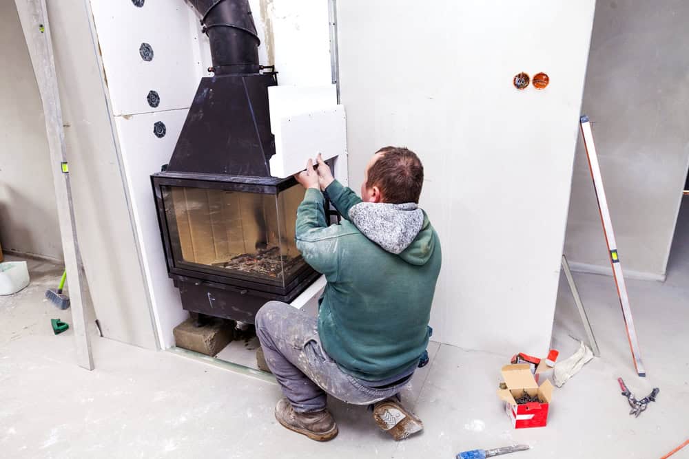 A person in work clothes kneels on the floor, installing drywall around a black fireplace insert—tools and materials scattered nearby, as part of a Chimney Repair Providence County, RI project in a room under construction.
