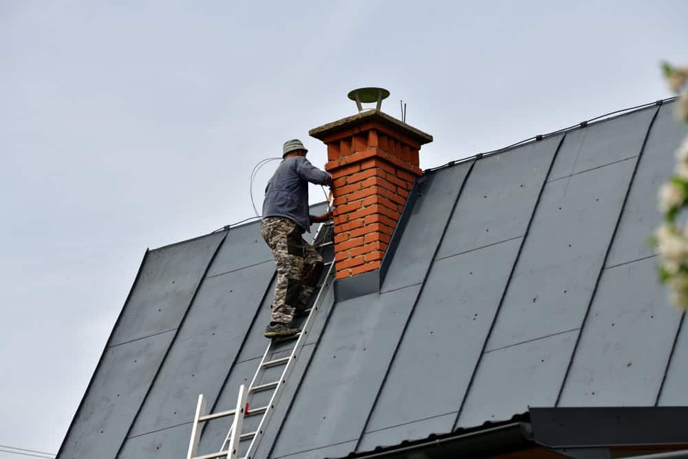 A person in camouflage pants and a jacket stands on a ladder, working on chimney repair atop a red brick chimney of a slanted, metal-roofed building under the clear Providence County, RI sky.