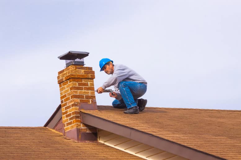 A worker wearing a blue hard hat and gray sweatshirt crouches on a roof in RI, inspecting or repairing a brick chimney with a tool in hand—an example of Chimney Cleaning Providence County under an overcast sky.