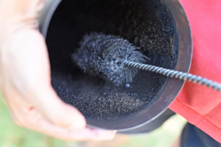 A close-up of a person cleaning the inside of a black metal pipe or cylinder with a wire brush, with black soot or residue visible inside the pipe.