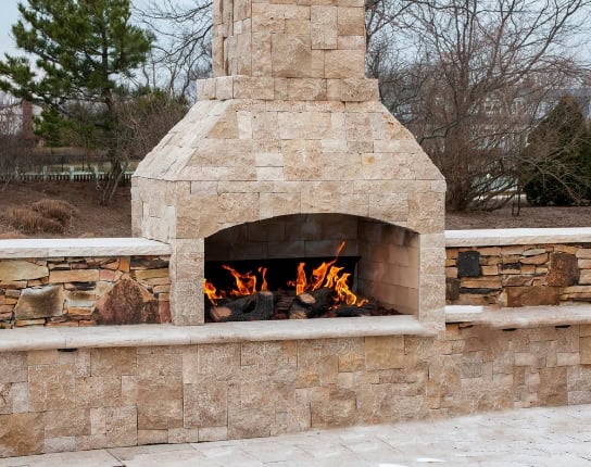A stone outdoor fireplace with a burning fire stands on a stone patio beside a stone wall, perfect for cozy gatherings. Surrounded by leafless trees and shrubs, it’s ideal for enjoying cool evenings—just remember to schedule a Chimney Sweep Providence County, RI.
