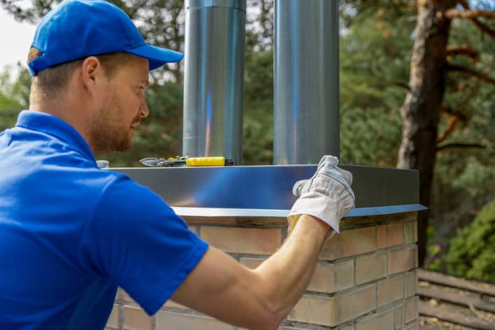 A worker in a blue uniform and cap installs or repairs a metal chimney cap on a brick structure outdoors, using tools and protective gloves. Chimney Repair Providence County, RI services ensure safe and efficient chimneys amid scenic trees.