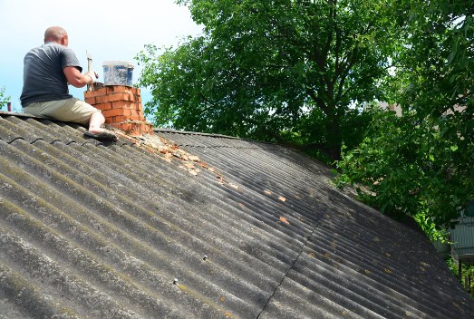 A person sits on a sloped corrugated roof in RI, repairing a red brick chimney with mortar and scattered bricks around, while green trees fill the background.