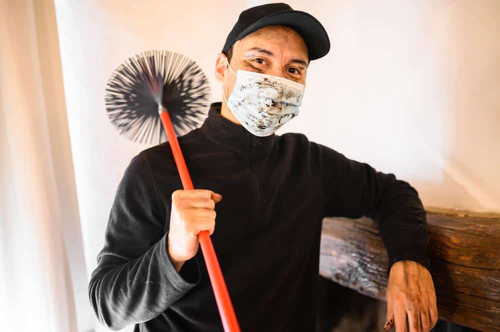 A person wearing a face mask, black cap, and black sweater stands indoors, leaning on a wooden mantel while holding a chimney sweep brush over their shoulder, ready for Chimney Sweep Providence County, RI services.