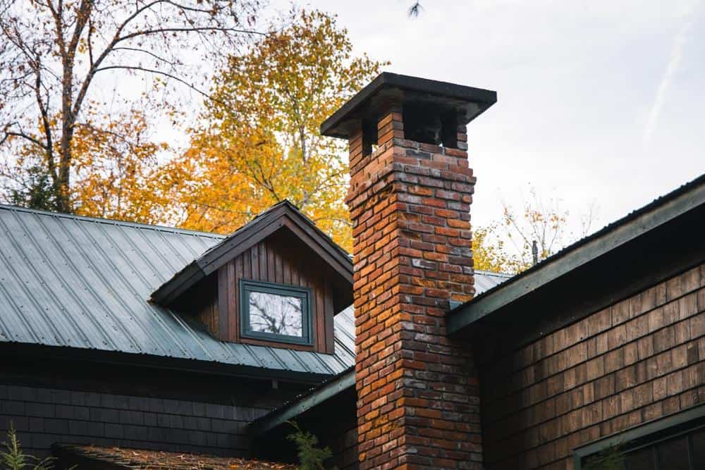 A brick chimney rises from the roof of a wooden house with a metal roof and a small dormer window, surrounded by trees with autumn foliage—perfect for those needing Chimney Repair Providence County services.