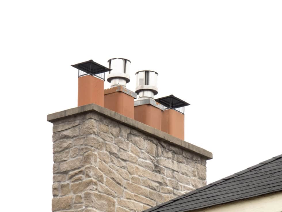 A stone chimney with four terracotta flue pipes, featuring mesh guards and stainless steel chimney cowls, stands on a rooftop against a white sky—typical of homes needing Chimney Repair Providence County or a Chimney Sweep Providence County in RI.