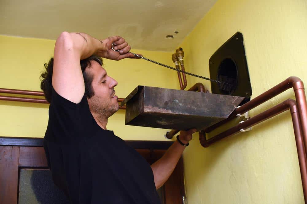 A man is cleaning a ventilation duct with a long brush in a room with yellow walls and copper pipes, similar to the thorough care seen in Chimney Cleaning Providence County, RI. He stands on a stool or ladder to reach the vent.