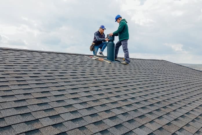 Two workers in blue caps and jackets install or repair a roof vent on a shingled roof under a partly cloudy sky. Tools and materials are nearby, showcasing the expertise needed for Chimney Cleaning Providence County, RI projects.
