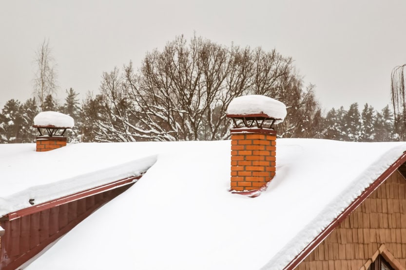 A snow-covered roof in RI with two brick chimneys, each topped with a thick layer of snow. Bare trees and a cloudy sky form the backdrop—a classic winter scene reminiscent of a Chimney Sweep Providence County postcard.