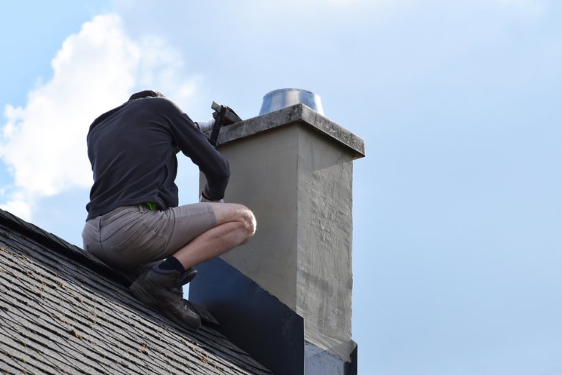 A person in shorts and a long-sleeve shirt kneels on a sloped roof, performing Chimney Repair Providence County, RI, with tools. The sky is partly cloudy in the background.