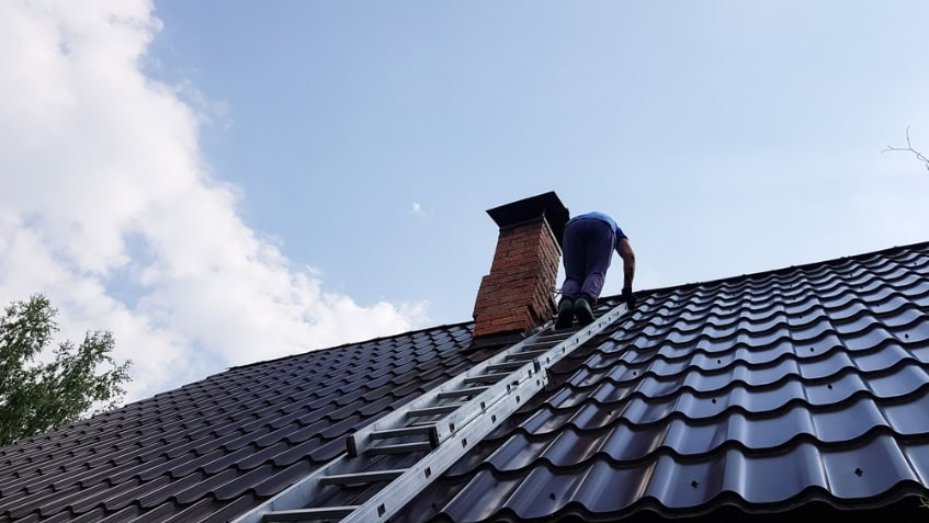 A person climbs a metal ladder on a wavy metal roof in RI, heading toward a brick chimney under a blue sky, possibly for Chimney Repair Providence County.