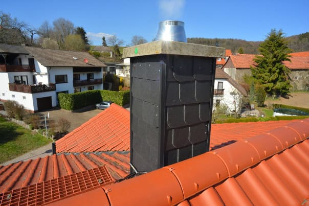A black chimney with a metal cap stands atop a red-tiled roof in RI, gently emitting white smoke. Nearby residential buildings, trees, and a clear blue sky complete the scene—ideal for those seeking Chimney Sweep Providence County services.