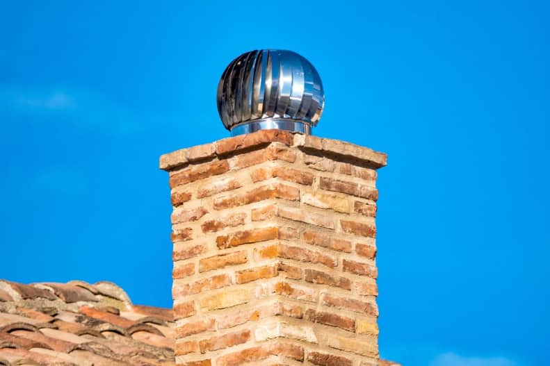 A brick chimney with a shiny, metallic rotating chimney cap stands against a clear blue sky, hinting at professional Chimney Sweep Providence County services. Part of a tiled roof is visible in the foreground.