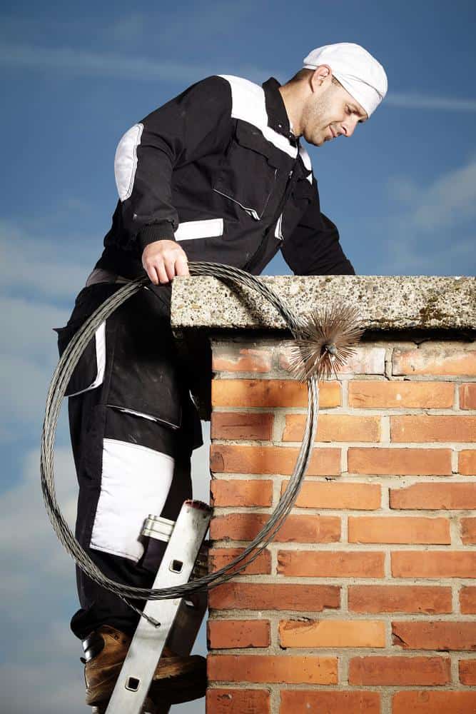 A chimney sweep in work clothes and cap stands on a ladder beside a brick chimney in RI, holding a flexible chimney brush and cable, under a blue sky.