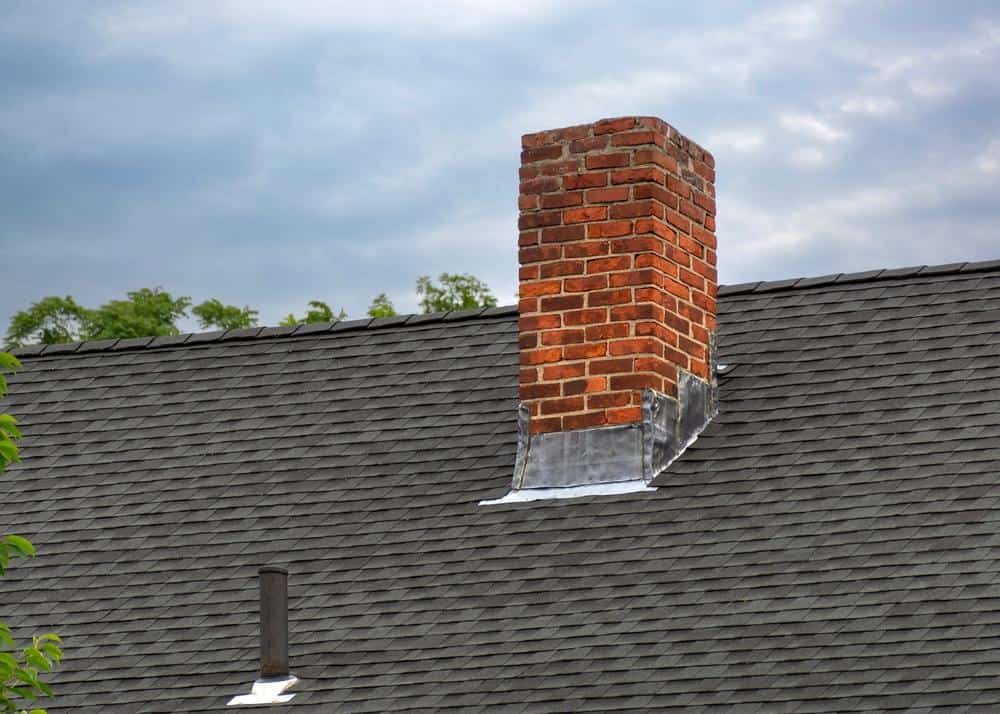 A red brick chimney with metal flashing stands on a dark gray shingled roof under a cloudy sky, with trees visible in the background—ideal for Chimney Cleaning Providence County, RI.
