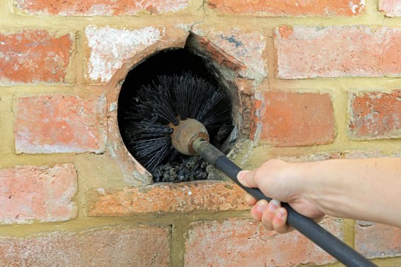 A person’s hand holds a chimney brush, cleaning the inside of a round opening in a brick wall—expert Chimney Cleaning Providence County, RI ensures your fireplace stays safe and efficient.