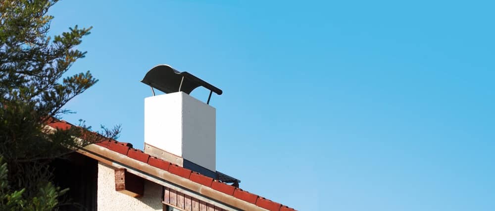 A white chimney with a black, curved chimney cap on a house with a red-tiled roof stands next to a leafy tree under a clear blue sky—perfect inspiration for anyone seeking Chimney Repair Providence County, RI.
