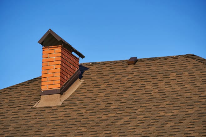 A close-up view of a house roof with brown shingles and a red brick chimney, ideal for Chimney Repair Providence County, set against a clear blue sky.