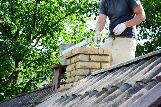 A person in gloves is performing Chimney Repair in Providence County, RI, working on a brick chimney atop a sloped roof, with trees and lush green foliage in the background.
