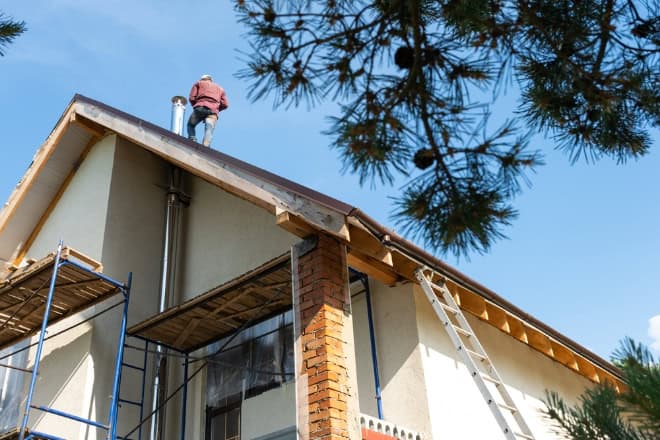 A person stands on the roof of a house under construction or repair, possibly preparing for Chimney Cleaning Providence County, RI, with scaffolding and a ladder set up alongside the building, surrounded by trees and a clear blue sky.