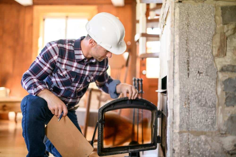 A person wearing a white hard hat and plaid shirt inspects a fireplace, holding a clipboard and opening the stove door inside a cozy, wood-accented home—a Chimney Sweep Providence County, RI professional at work.
