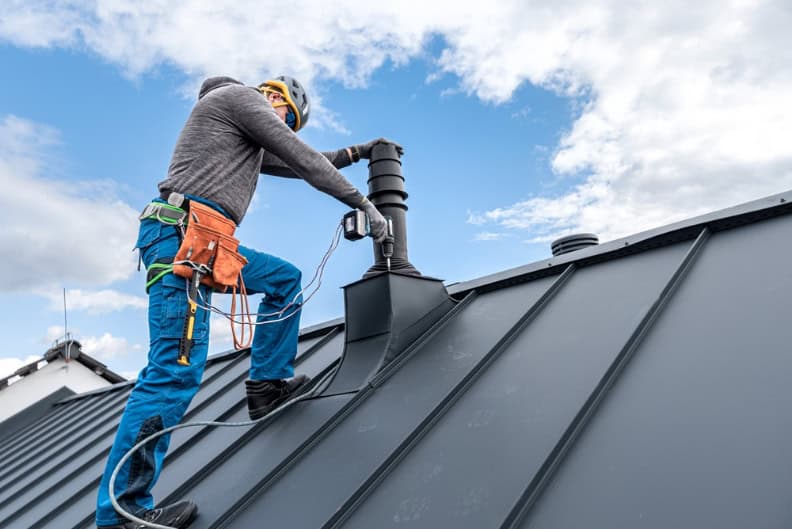 A worker in safety gear stands on a sloped metal roof in RI, using power tools to install or repair a chimney vent under a partly cloudy sky. Tool belt and harness visible for safety.