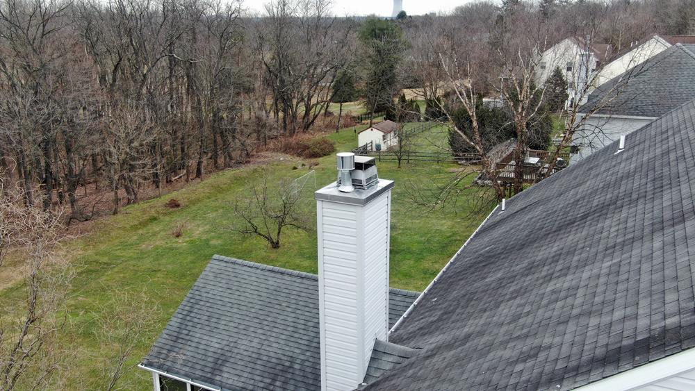 A rooftop with a white chimney rises above a gray-shingled RI home. The backyard features green grass, scattered trees without leaves, and a wooded area in the distance under an overcast sky—perfect for a Chimney Sweep Providence County recommends.