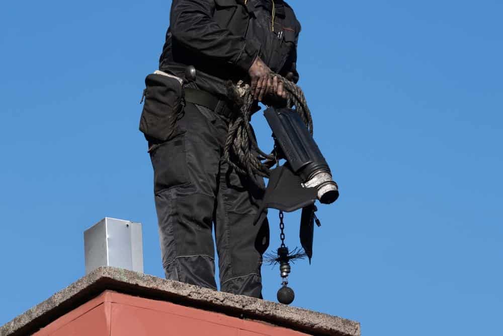 A person in dark work clothes stands on a rooftop holding chimney cleaning equipment, including a brush with a weighted ball, against a clear blue sky—perfectly capturing Chimney Sweep Providence County, RI in action.