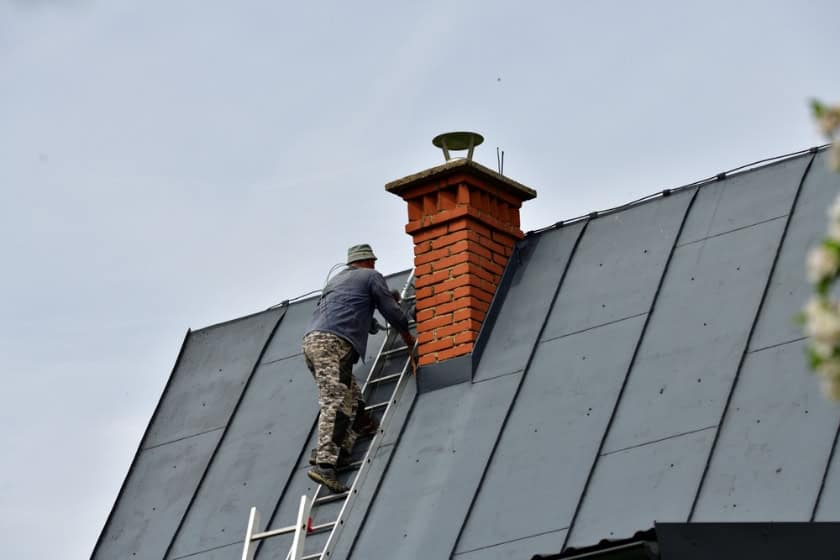 A person in camouflage pants climbs a ladder propped against a sloped metal roof to inspect or perform Chimney Cleaning Providence County, RI, on a red brick chimney under a cloudy sky.