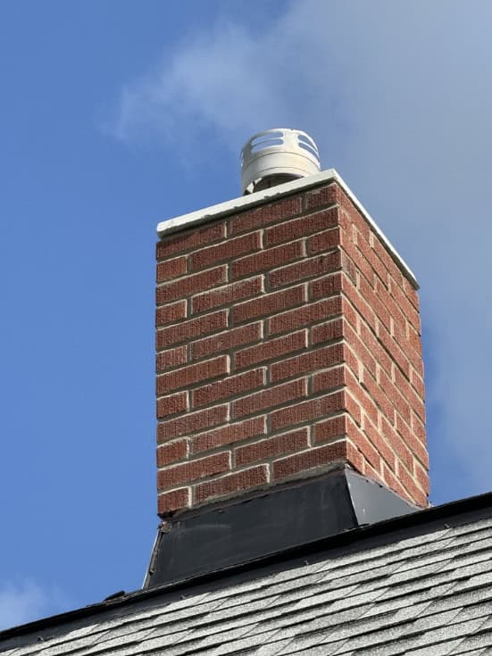 A red brick chimney with a metal cap extends from a gray shingled roof against a blue sky with some clouds, showcasing expert Chimney Repair Providence County, RI workmanship.