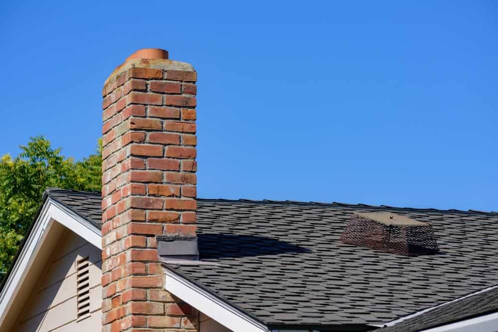 A close-up of a house roof with dark shingles, a red brick chimney, and a metal chimney cap against a clear blue sky in RI. Some green tree foliage is visible in the background—a great example for Chimney Repair Providence County, RI.