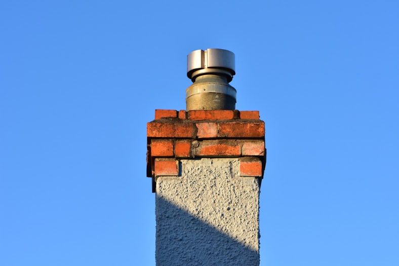 A close-up of a brick and stucco chimney with a metal cap, set against a clear blue sky. Sunlight casts a shadow on the lower part—perfect inspiration for anyone seeking Chimney Cleaning Providence County, RI.