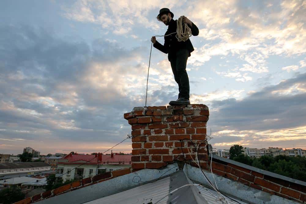 A person in dark clothing and a hat stands atop a brick chimney on a rooftop, holding a rope, with a cloudy sky and city buildings in the background.